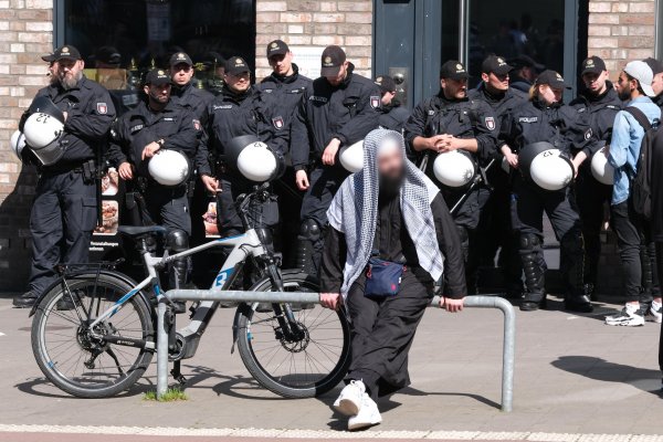 Auf dem Bild ist ein junger Muslim zu sehen. Er lehnt an einem Geländer am Rande des Bürgersteiges. Hinter ihm steht eine große Gruppe uniformierter Polizisten.