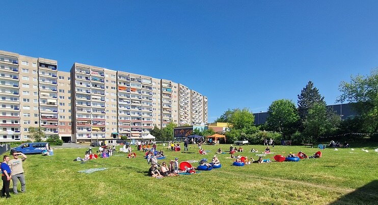 viele Personen auf einer Wiese sitzend im Hintergrund ein Stadthaus